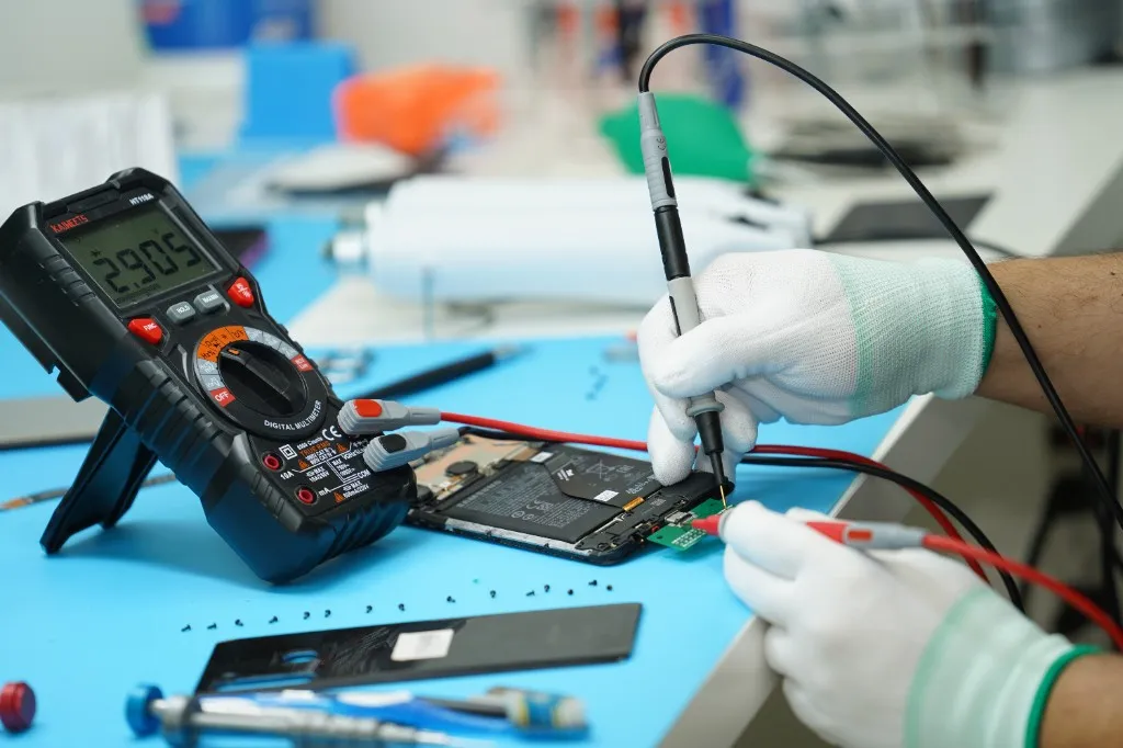 Technician testing a disassembled phone with a multimeter on a repair bench