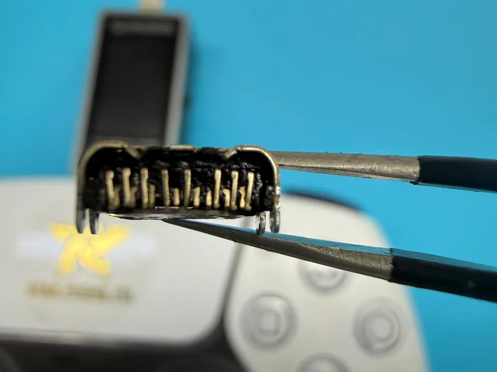 Puerto de carga USB-C quemado retirado de un control DualSense de PS5, sostenido con pinzas