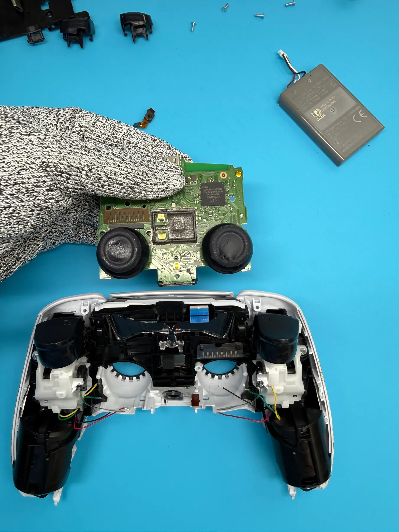Technician's gloved hand repairing a disassembled PlayStation controller's circuit board and joysticks