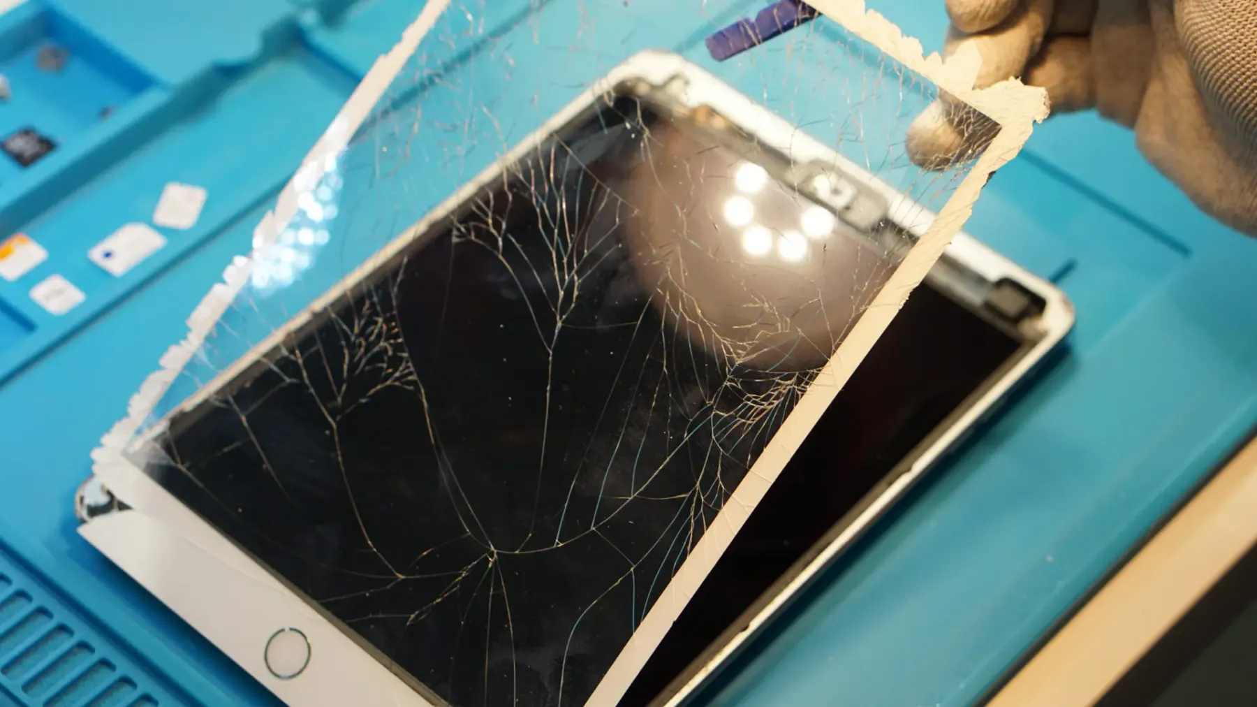 Technician lifting a shattered tablet screen during repair under a ring light