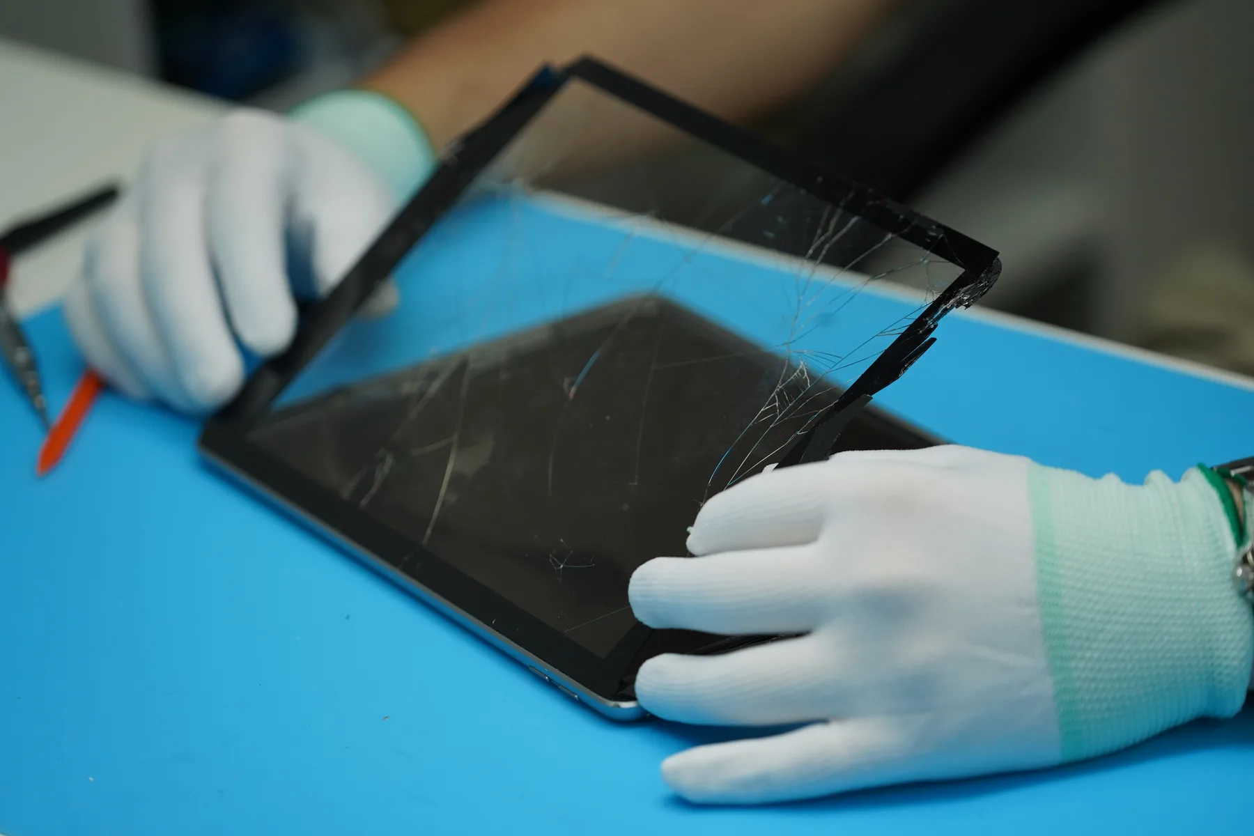 Technician lifting a shattered tablet screen during repair under a ring light